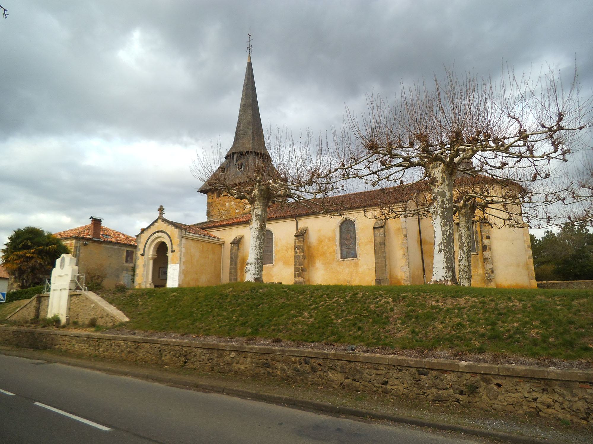 Eglise de Laurède. Elle se distingue par son clocher en flèche, ses murs aux teintes jaunes et ocres, et des fenêtres en arc typiques des églises traditionnelles. Au premier plan, des arbres taillés sans feuilles bordent le terrain surélevé.