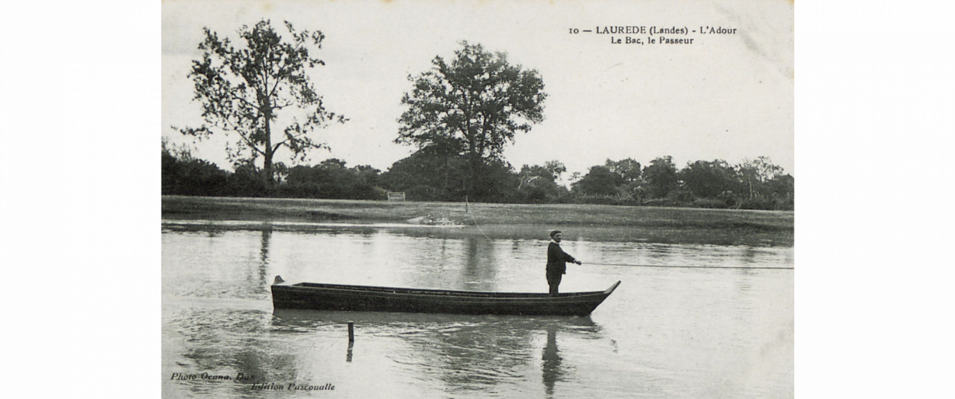 Photo en noir et blanc du bac sur l'Adour. Seul le passeur est sur le bac. Pour traverser, il se guide grâce à une corde tendue entre les deux rives.